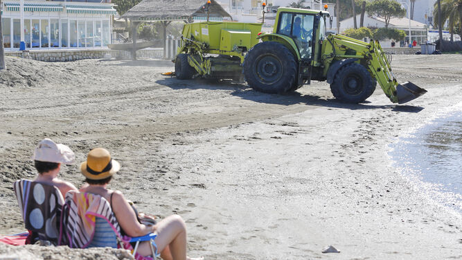 Una pareja toma el sol en la playa de Pedregalejo mientras una máquina trabaja en su arreglo.