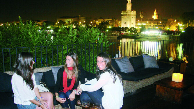 Tres jóvenes disfrutan de la noche sevillana en una terraza junto al río y con la Torre del Oro de fondo.