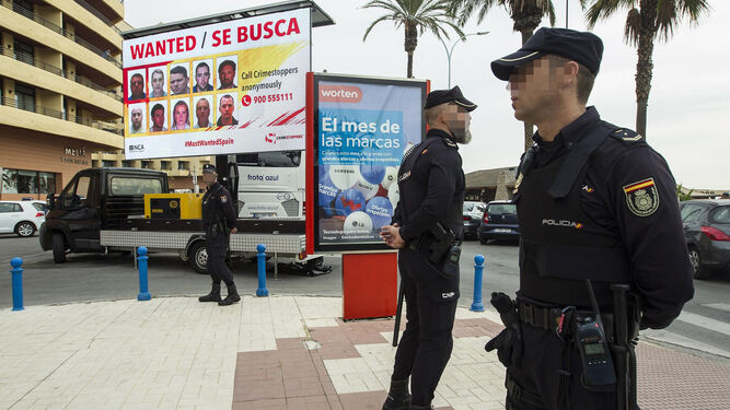 Dos policías nacionales junto a un cartel con la imagen de varios fugitivos.