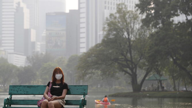 Una mujer sentada en el Parque Lumpini en Bangkok junto a edificios de gran altura, envueltos en  'smog' y neblina de polvo fino pesado.